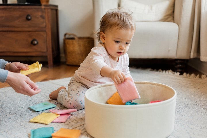 A 14-month-old posting soft fabric squares into a wide container with a parent nearby