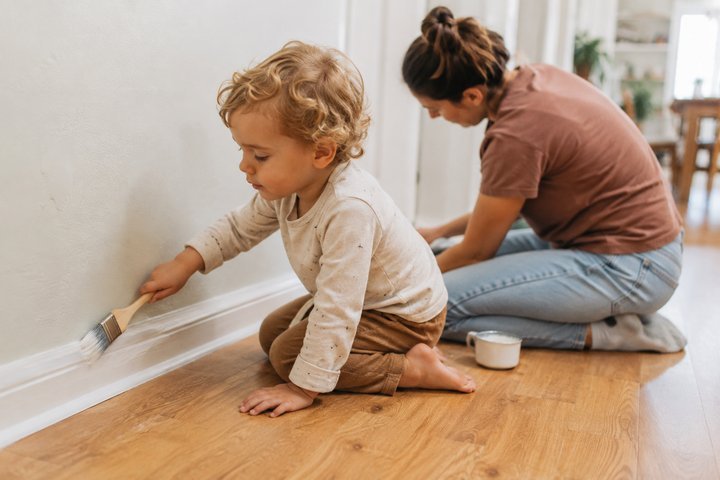 A 2-year-old using a clean dry paintbrush along a baseboard while a parent works nearby