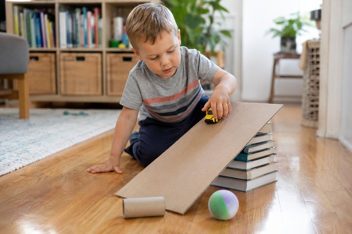 A 3-year-old testing a toy car on a homemade cardboard ramp propped on a stack of books