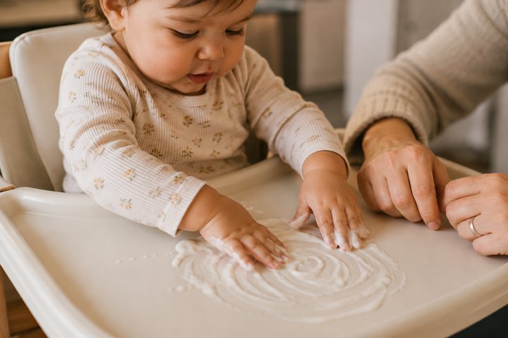 Close-up of a 1-year-old making finger trails through plain yogurt on a high-chair tray with a parent's hands beside them